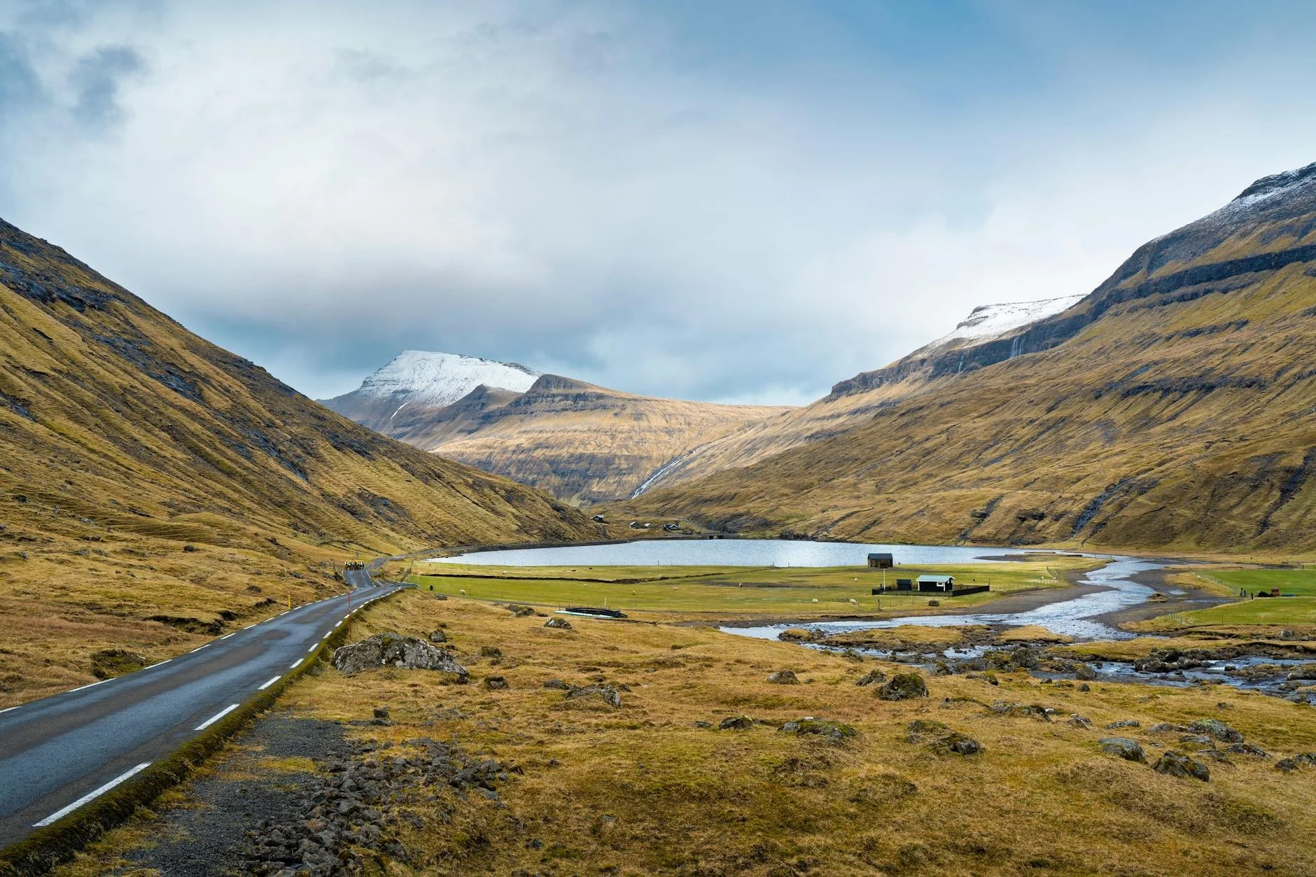 Picturesque valley in Faroe Islands showcasing snowy mountain peaks, a winding road, and serene landscape.